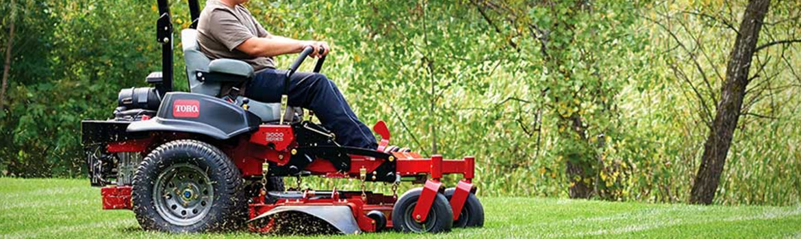 A person using a zero-turn lawn mower on a field of green grass near a row of trees during the day.